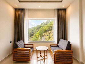 Una habitación con un ventanal grande, dos sillas de madera, una mesa redonda y vistas a una ladera boscosa.