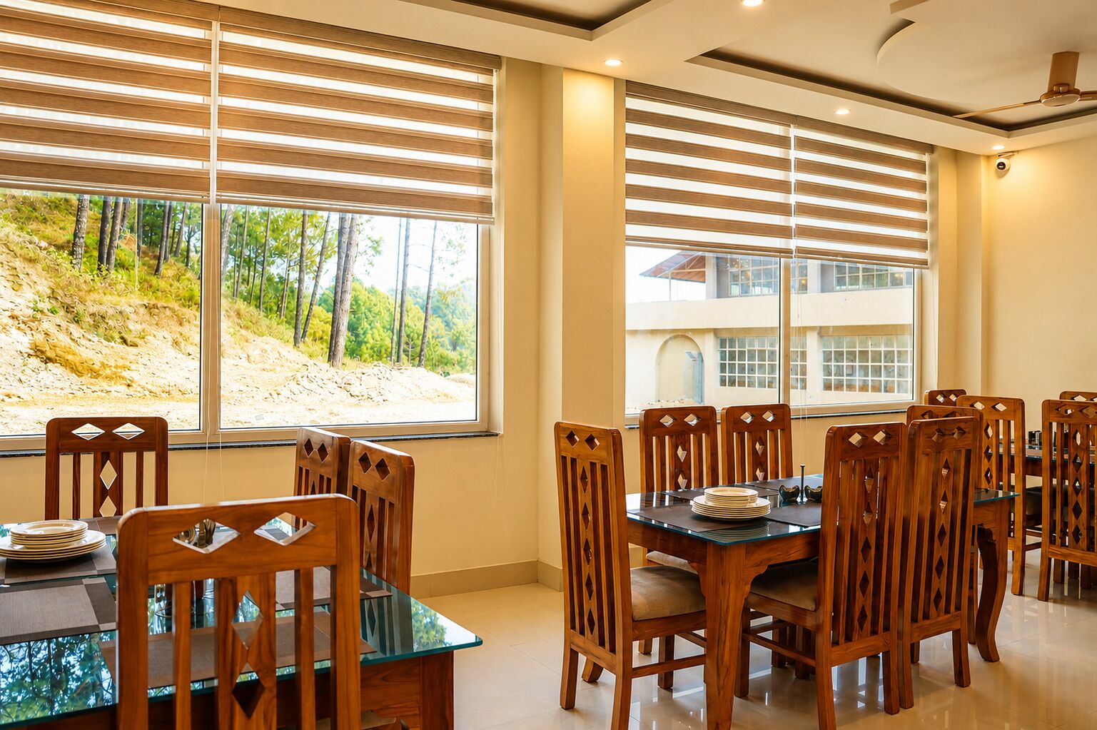 A dining area with a glass table and wooden chairs, large windows with blinds, and a view of a hillside.