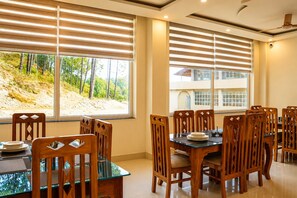 Un comedor con mesa de vidrio y sillas de madera, ventanas grandes con persianas y vista a una ladera.