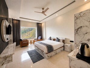 Habitación de hotel moderna con una cama grande, un escritorio, una silla, un televisor montado en la pared y vistas a un bosque por la ventana.