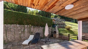 Un patio al aire libre con una mesa de madera y dos sillones, un muro de piedra y una vista de una casa al fondo.