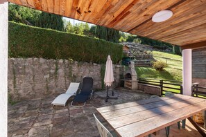 Un patio al aire libre con una mesa de madera y dos sillones, un muro de piedra y una vista de una casa al fondo.