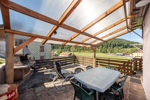 An outdoor patio with a table and chairs, a grill, and a view of a green landscape.