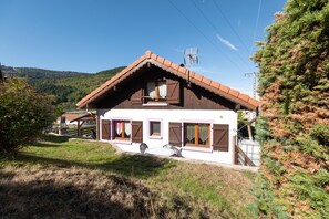 A wooden chalet with a sloped roof, a balcony, and a view of a forested hillside.