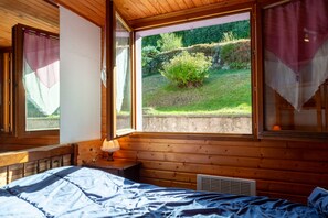 A wooden cabin room with a bed, a nightstand, a lamp, and a window overlooking a garden.