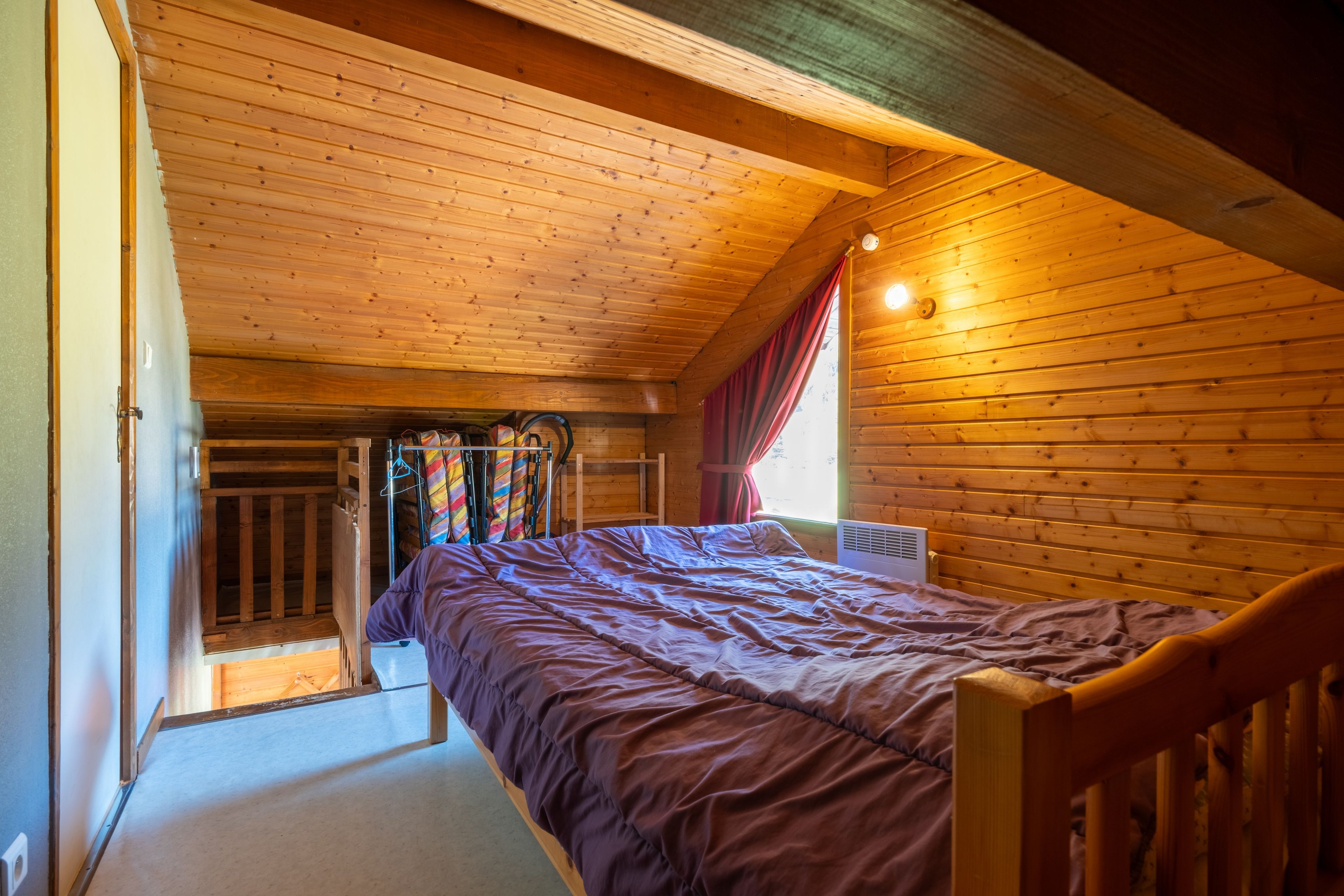 Une chambre avec un plafond en bois, un lit recouvert d’une courtepointe violette, une fenêtre avec des rideaux rouges et un radiateur.