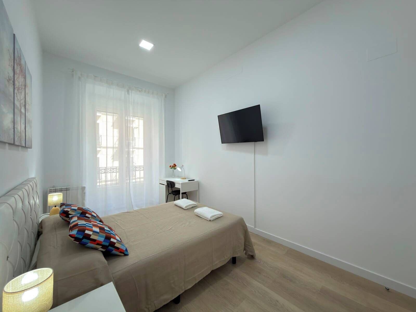 Habitación de hotel con cama, escritorio, silla, televisor montado en la pared y ventana con cortinas.