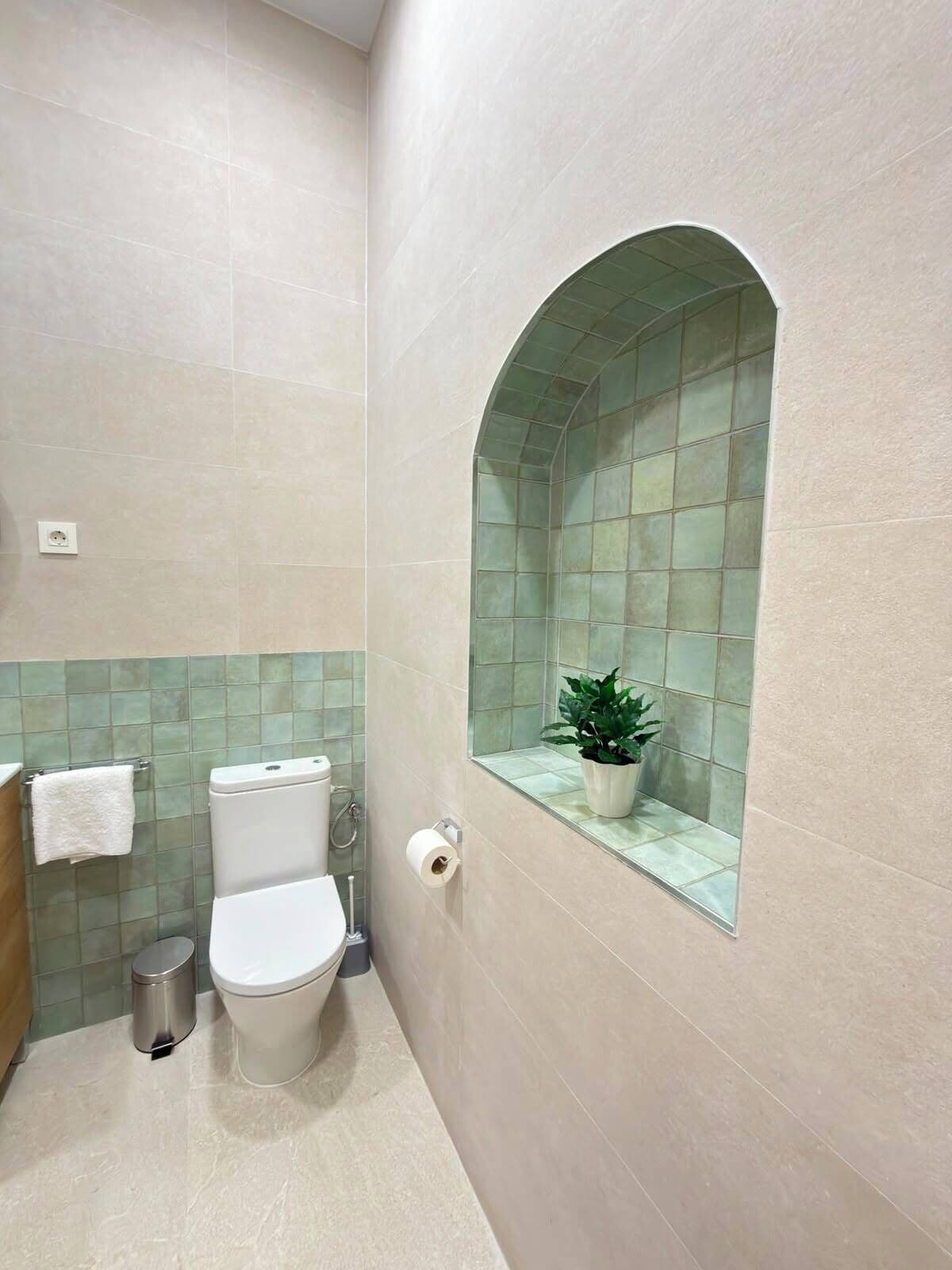 Een badkamer met toilet, prullenbak, handdoekhouder en een potplant.