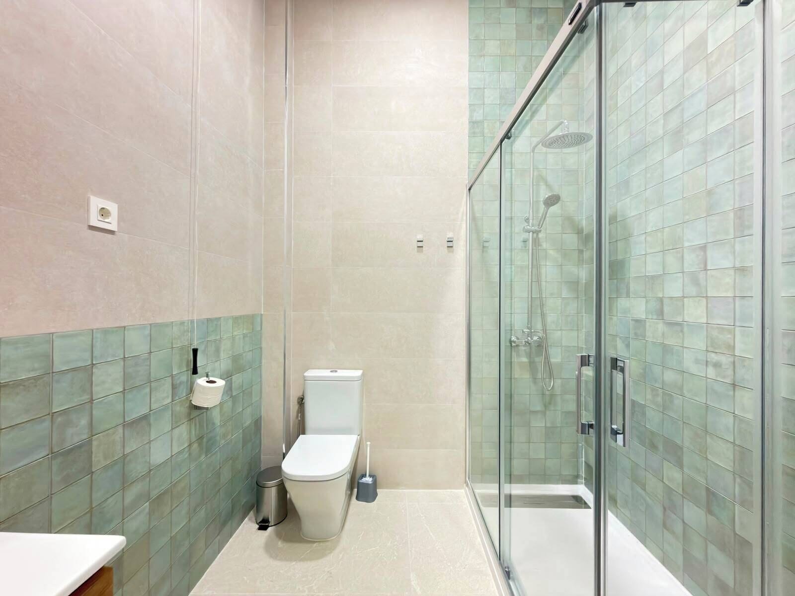 Un baño con inodoro, un cubo de basura y zona de ducha con mampara de vidrio.