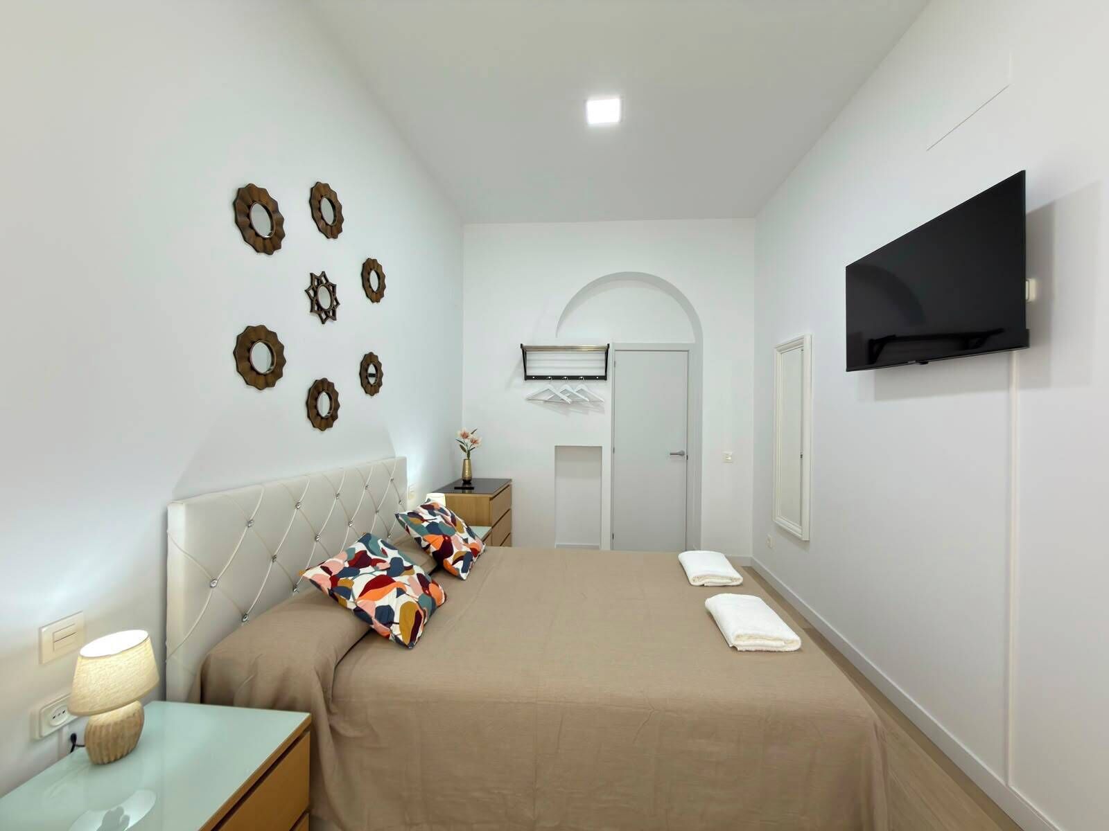 Un dormitorio con cama, mesita de noche, lámpara, televisor montado en la pared y espejos decorativos en la pared.
