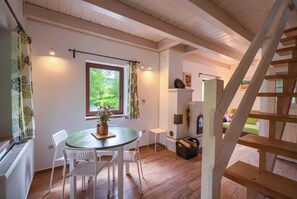 A cozy room with a wooden floor, a round table with chairs, a window with a view of greenery, and a wooden staircase.