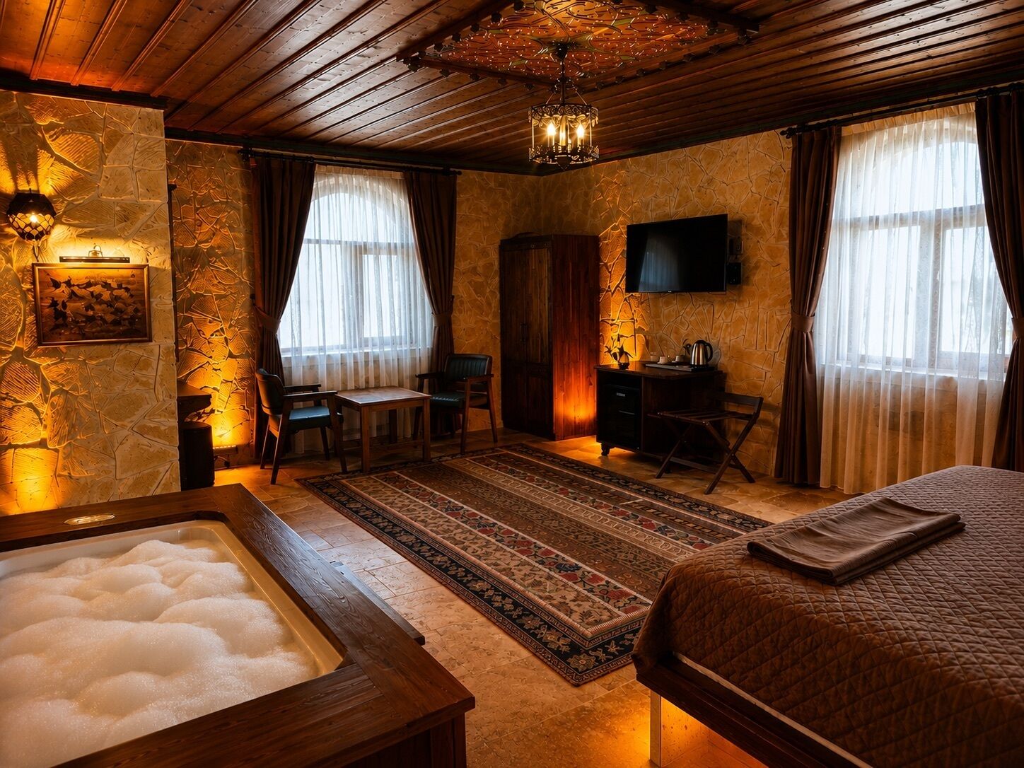 A room with a stone wall, a wooden ceiling, a bed, a desk, a chair, a television, and a hot tub.