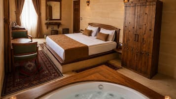 Habitación de hotel con una cama grande, un armario de madera, zona de estar con una silla, una mesita pequeña, baño con lavamanos y espejo, y un jacuzzi.