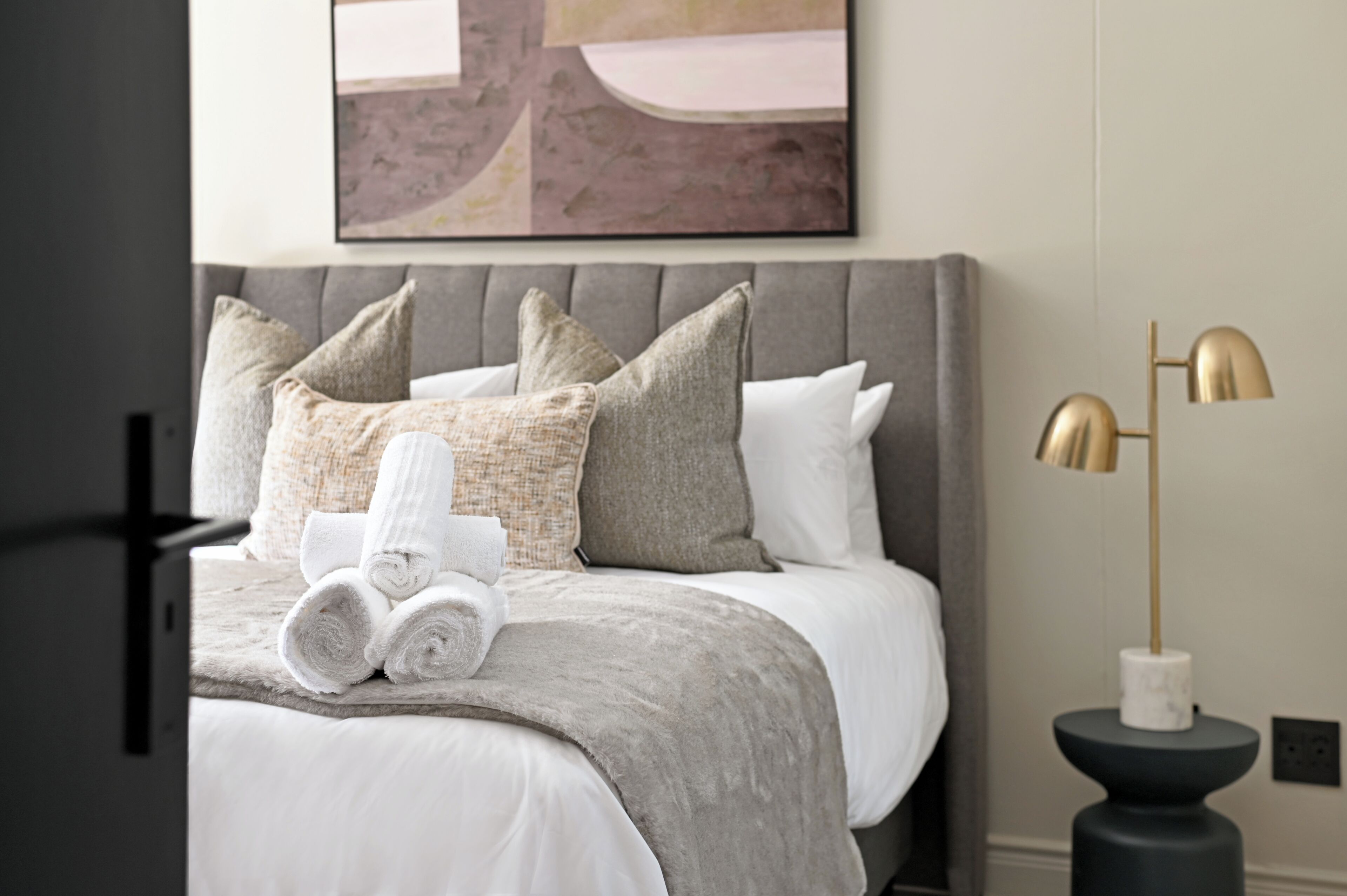 Una cama bien hecha con cabecera gris y ropa de cama blanca, un juego de toallas enrolladas y una lámpara de pie moderna al lado de la cama.