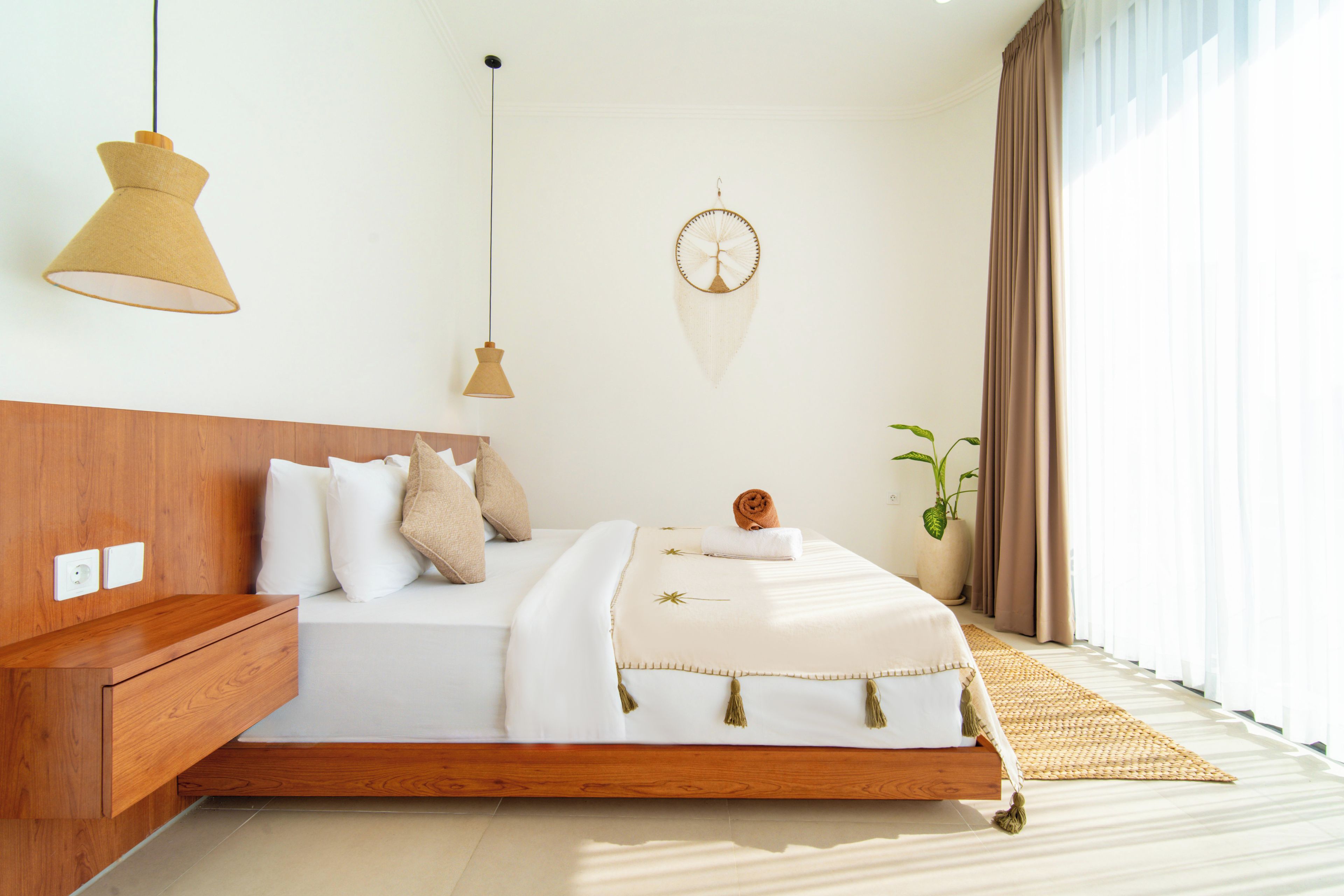 A modern bedroom with a wooden headboard, a bed with white bedding, a hanging light fixture, a dreamcatcher, and a potted plant.