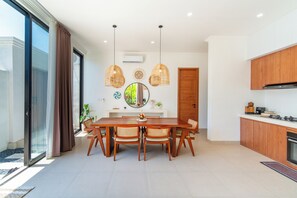 A modern kitchen with wooden cabinets, a dining table with chairs, and a large window with curtains. - 2-Min Walk to Seseh Beach • 2BR Private Pool Villa with Rooftop Sea View (Badung)