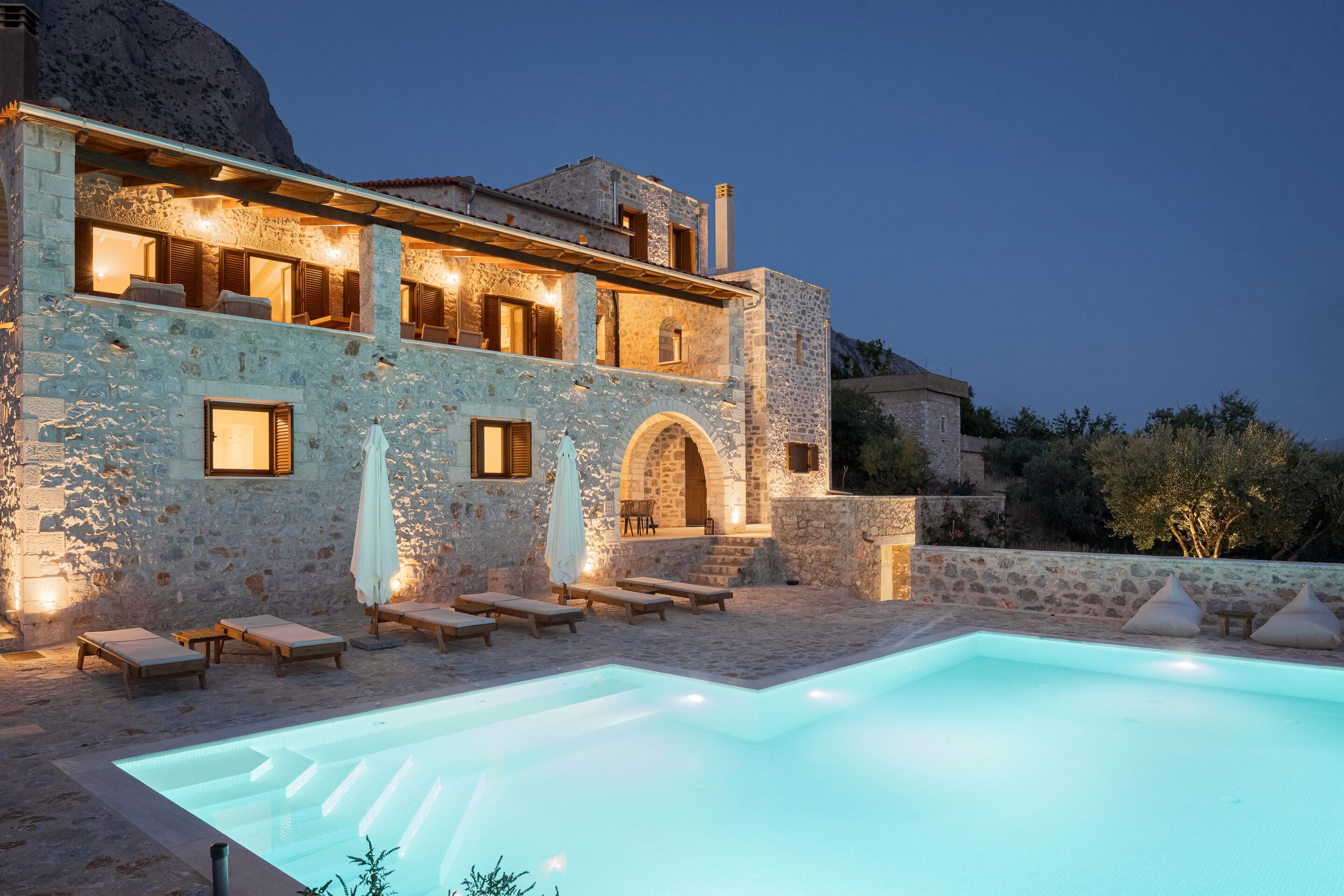 A stone building with a swimming pool, outdoor seating, and a mountainous backdrop.