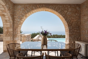 Un arc en pierre mène à une table dont le plateau est en verre, entourée de chaises en osier, et d’où l’on a vue sur une piscine et sur la végétation environnante.