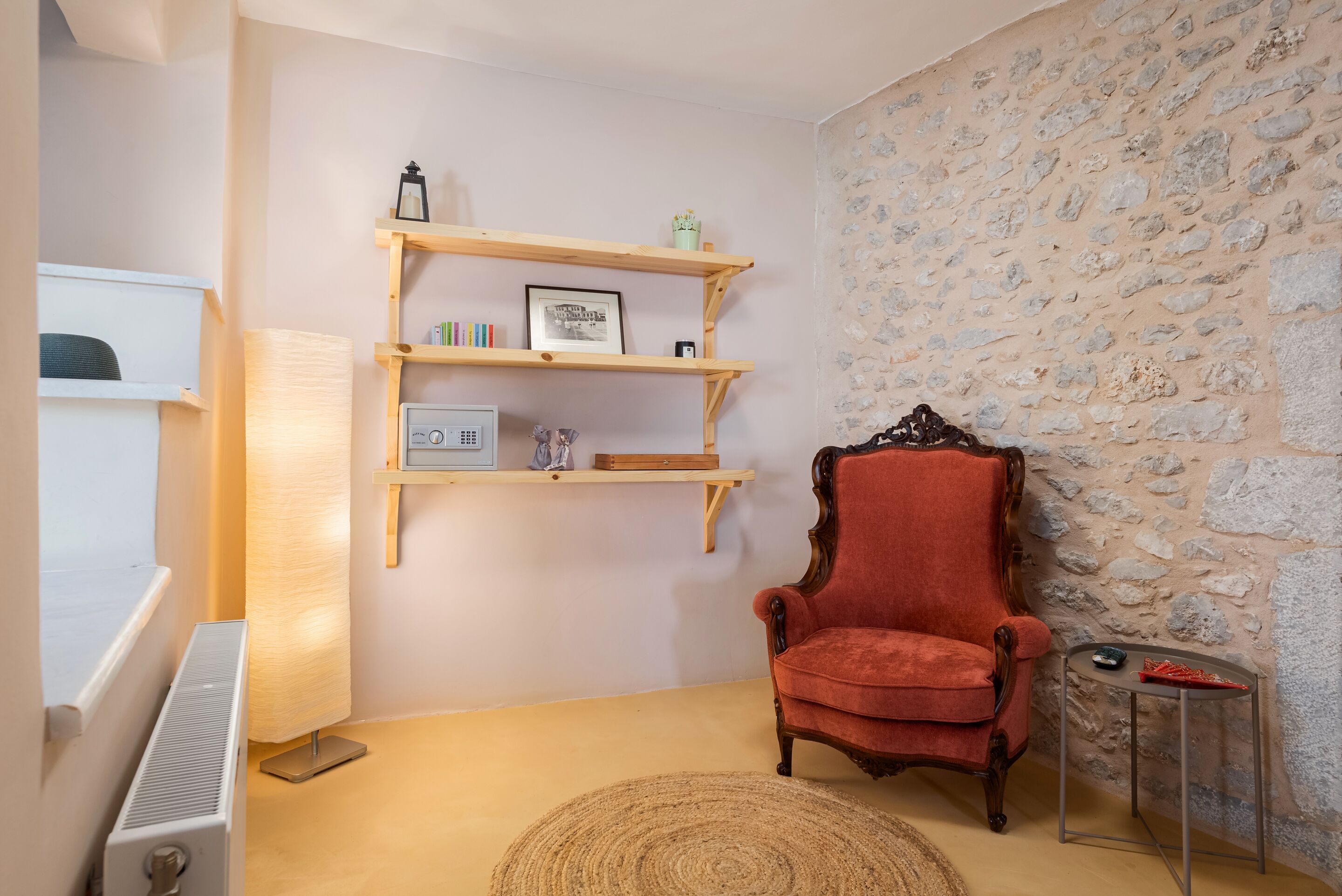 Une pièce avec un fauteuil rouge, un radiateur, une lampe, un tapis tissé et un mur en pierre.