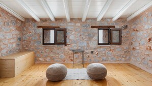 A room with stone walls, wooden beams, two windows, a small table, and two round ottomans. - Pirgos Petropoulea (Agios Nikon)