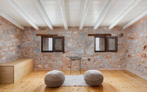 A room with stone walls, wooden beams, two windows, a small table, and two round ottomans.