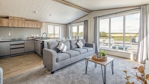 A modern living space with a grey sofa, a dining area, and large windows offering a view of the outdoors. - Lodge No 173 | Stewart's Resort Cameron (Cameron)