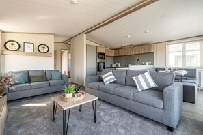 A modern living room with grey sofas, a coffee table, and a kitchen in the background. - Lodge No 173 | Hot Tub | Stewart's Resort Cameron (Cameron)