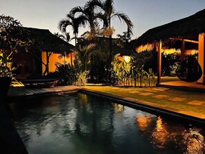 Ein Pool mit einem strohgedeckten Gebäude, Palmen und einem gut beleuchteten Gartenbereich.