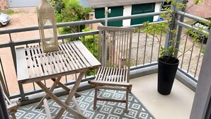 Een balkon met een houten tafel en stoel, een plant in pot en uitzicht op een groen gebouw en bomen.