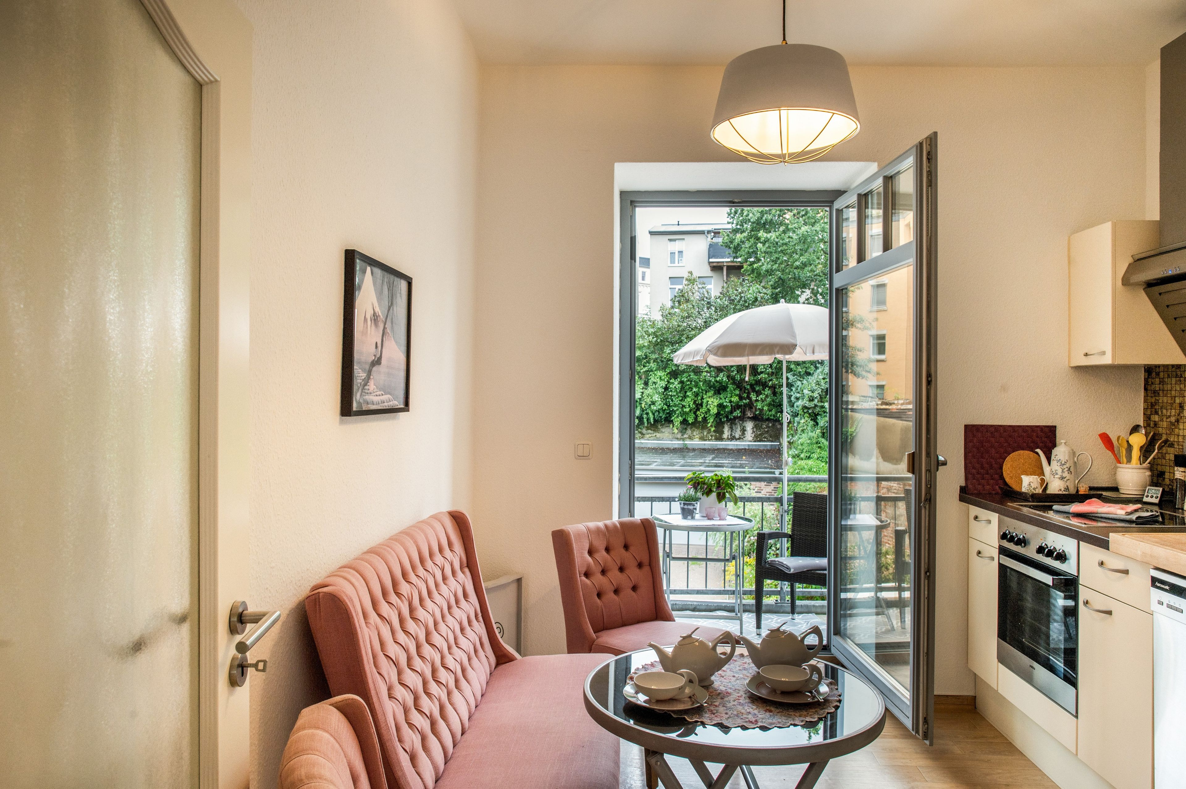 Cocina compacta con electrodomésticos modernos, comedor con mesa dispuesta para el té y vistas a un balcón con vegetación.