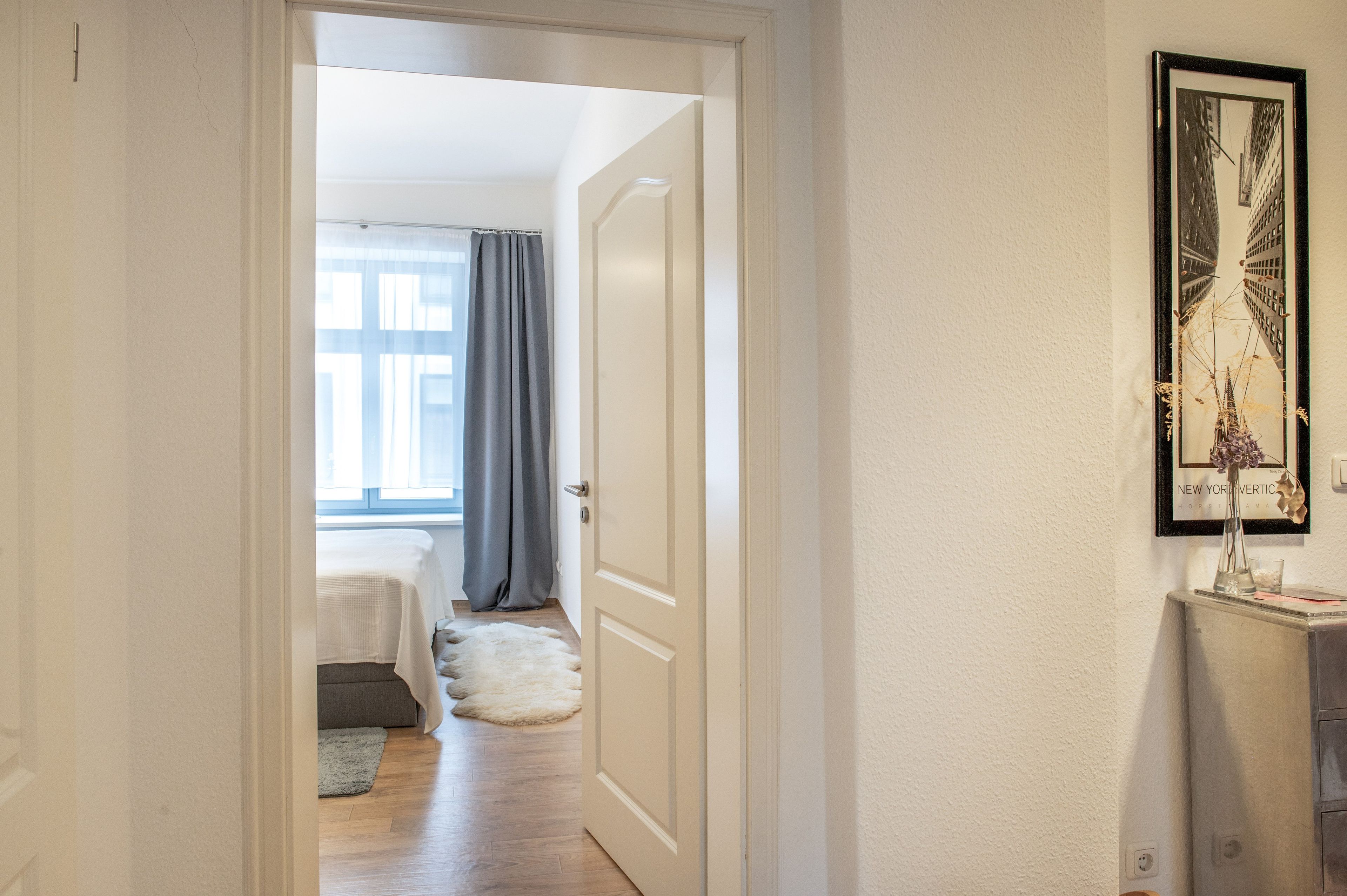 A hallway leading to a bedroom with a bed, a window with curtains, and a framed picture on the wall.