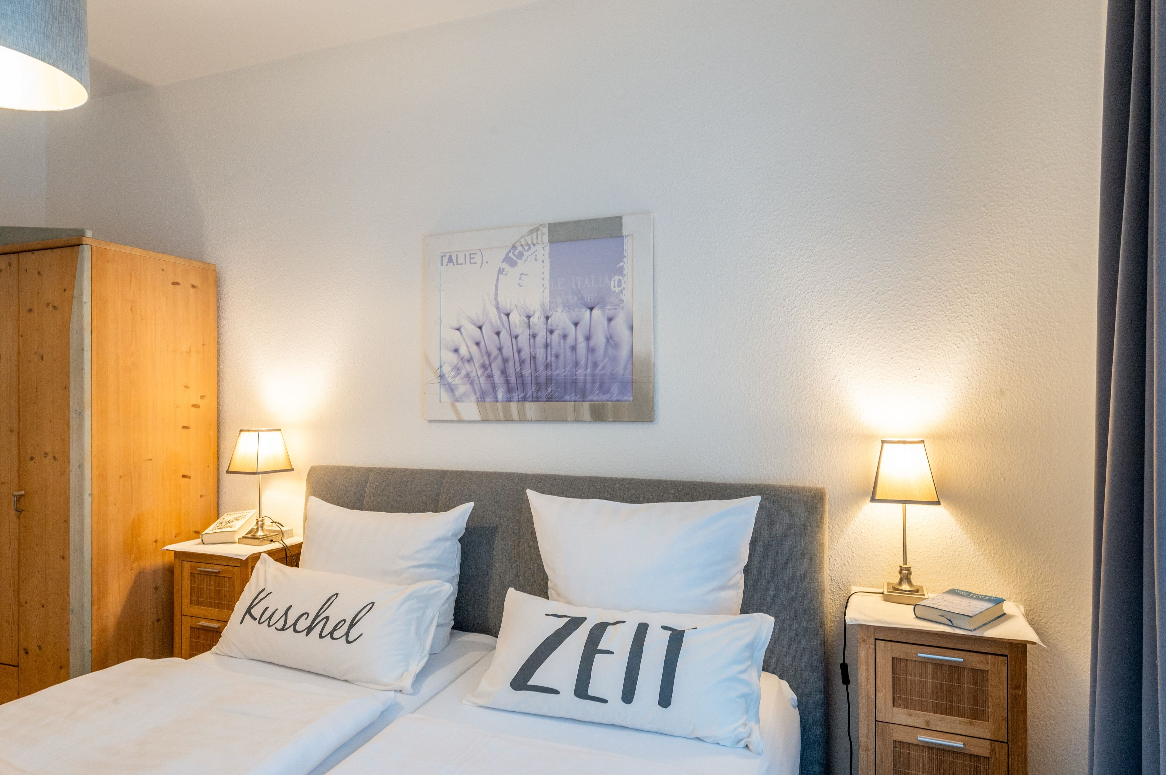 Hotellihuone, jossa on sänky, kaksi yöpöytälamppua, puinen vaatekaappi, seinällä oleva kehystetty taulu ja ikkuna, jossa on verhot.