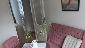 Een kamer met een roze bank, een bijpassende fauteuil, een klein rond tafeltje met een witte kan en roze accessoires, en een schilderij aan de muur.