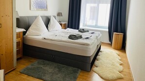 Een slaapkamer met een bed, kussens, een nachtkastje, een lamp, een raam met gordijnen, een houten garderobe en een vloerkleed.