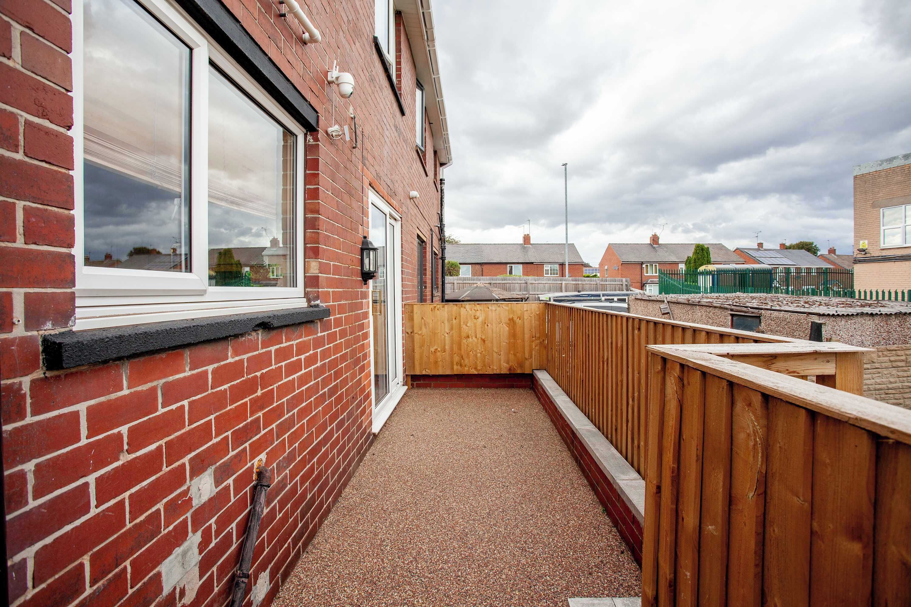 A narrow outdoor corridor with a red brick wall, a wooden fence, and a gravel path.