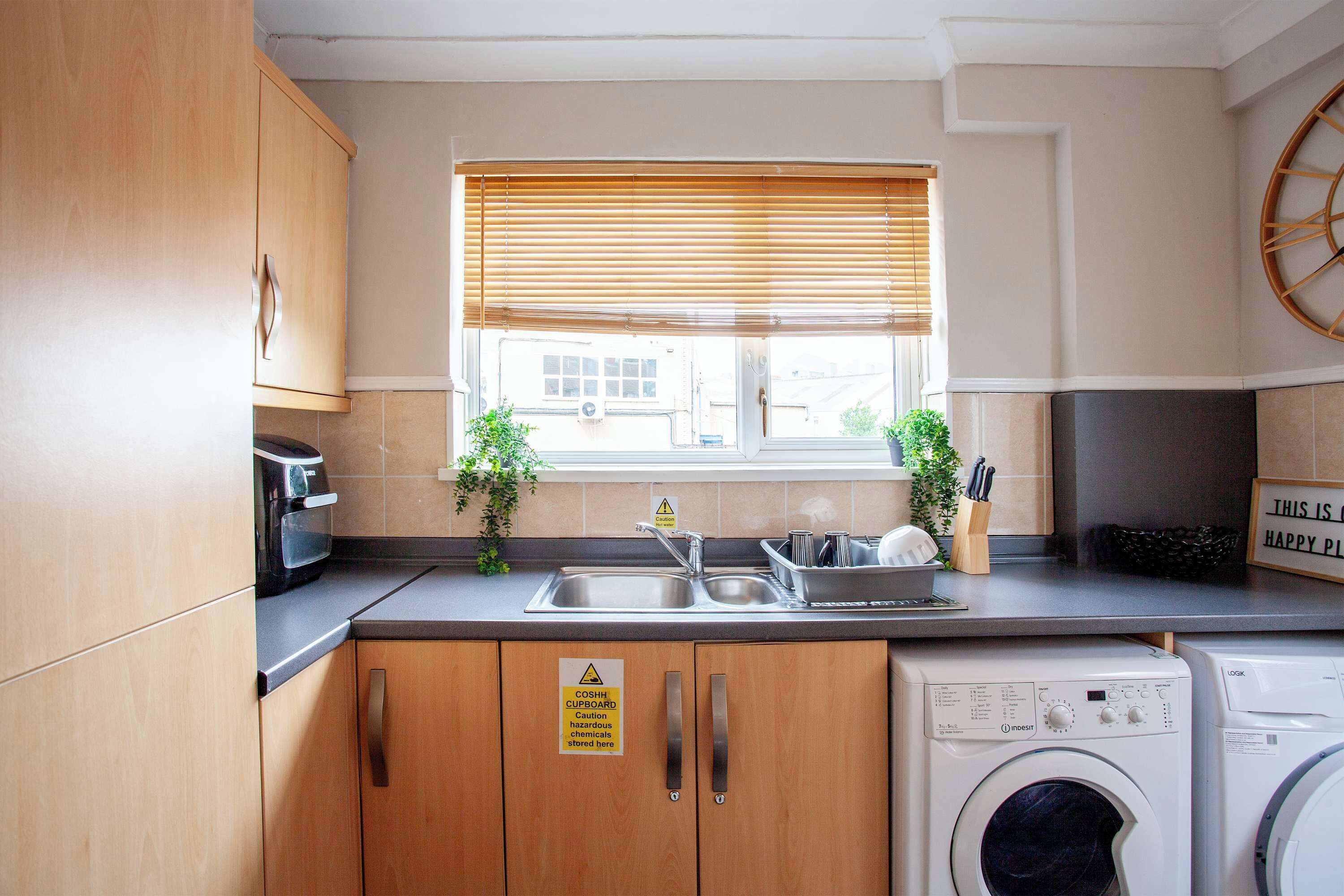 A compact kitchen with wooden cabinets, a stainless steel sink, a washing machine, and a window with blinds.