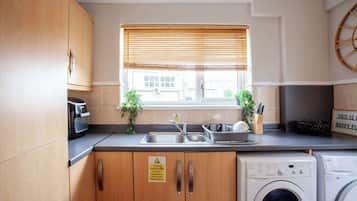 A compact kitchen with wooden cabinets, a stainless steel sink, a washing machine, and a window with blinds.