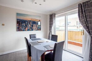 Un comedor con una mesa preparada para cuatro, un cuadro de paisaje urbano en la pared y vistas a un patio trasero a través de puertas corredizas de vidrio.