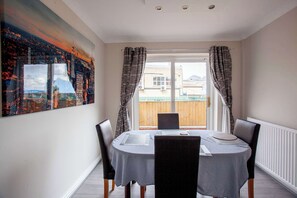 A dining area with a table set for four, a cityscape painting on the wall, and a view of a wooden fence through the window. - Peaceful Countryside Lodge: Scenic Views & (Barnsley)