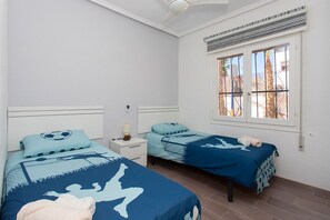 Ein Zimmer mit zwei Betten, jeweils mit blauer Bettdecke mit Silhouetten-Motiv, einem Nachttisch mit Lampe und einem Fenster mit Blick auf Palmen.