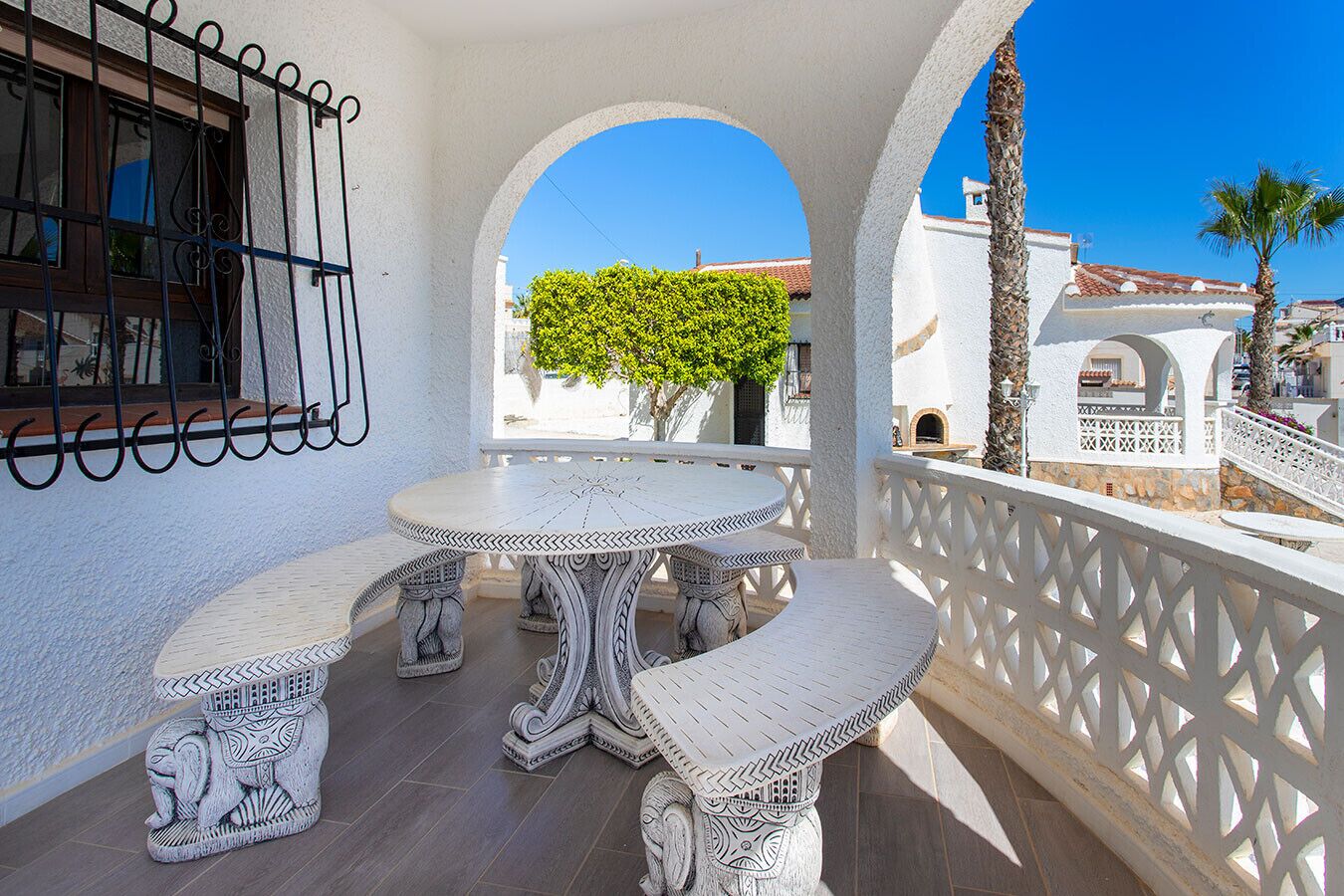Un balcon blanc avec une table ronde et des bancs, une balustrade blanche, et une vue sur un arbre et des immeubles.