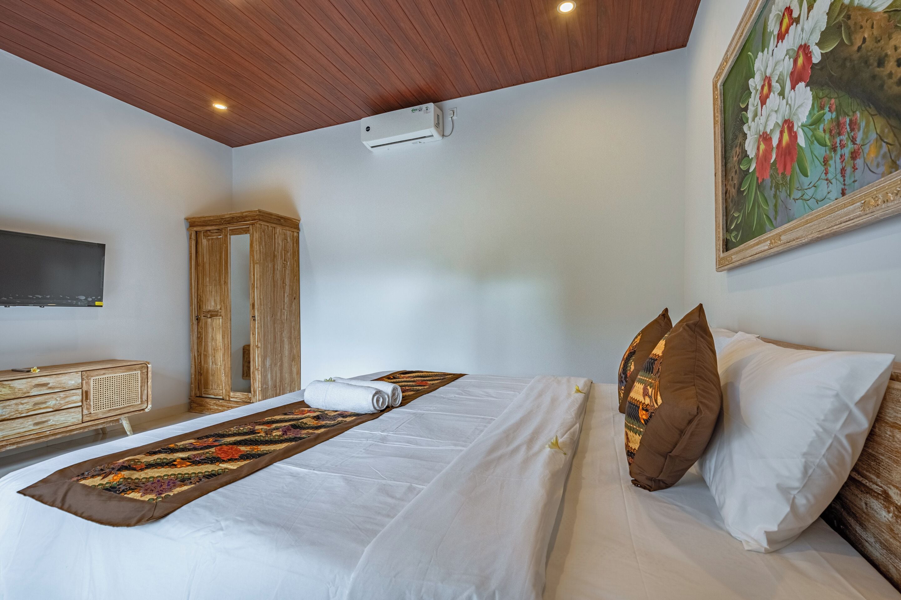A hotel room with a bed, a wooden wardrobe, a painting, and a ceiling with recessed lighting.
