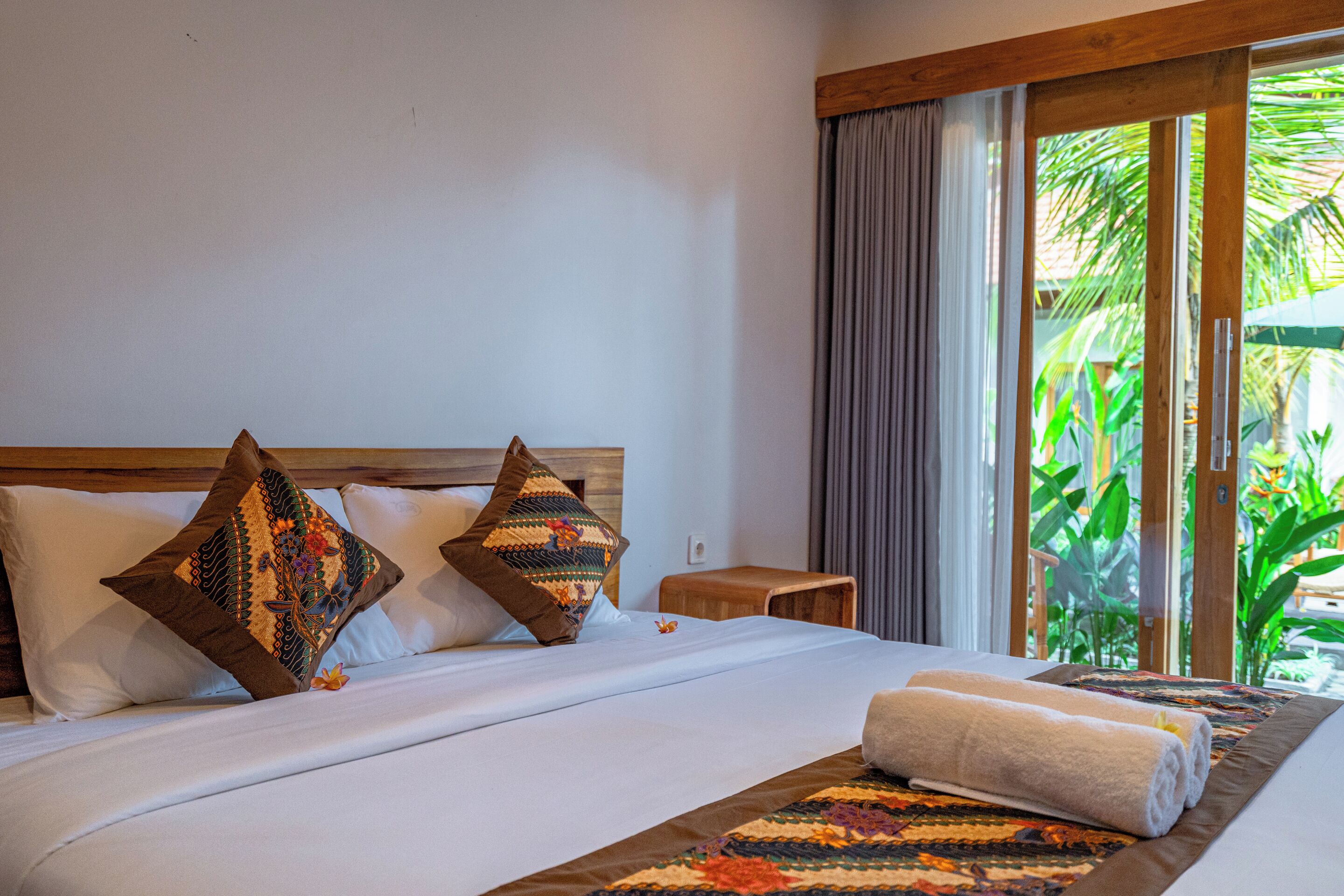 Habitación de hotel con cama, almohadas, una mesita de noche de madera y vistas a zonas verdes por la ventana.