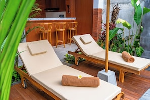 Una terraza de madera con dos sillones, una pequeña barra y plantas tropicales.