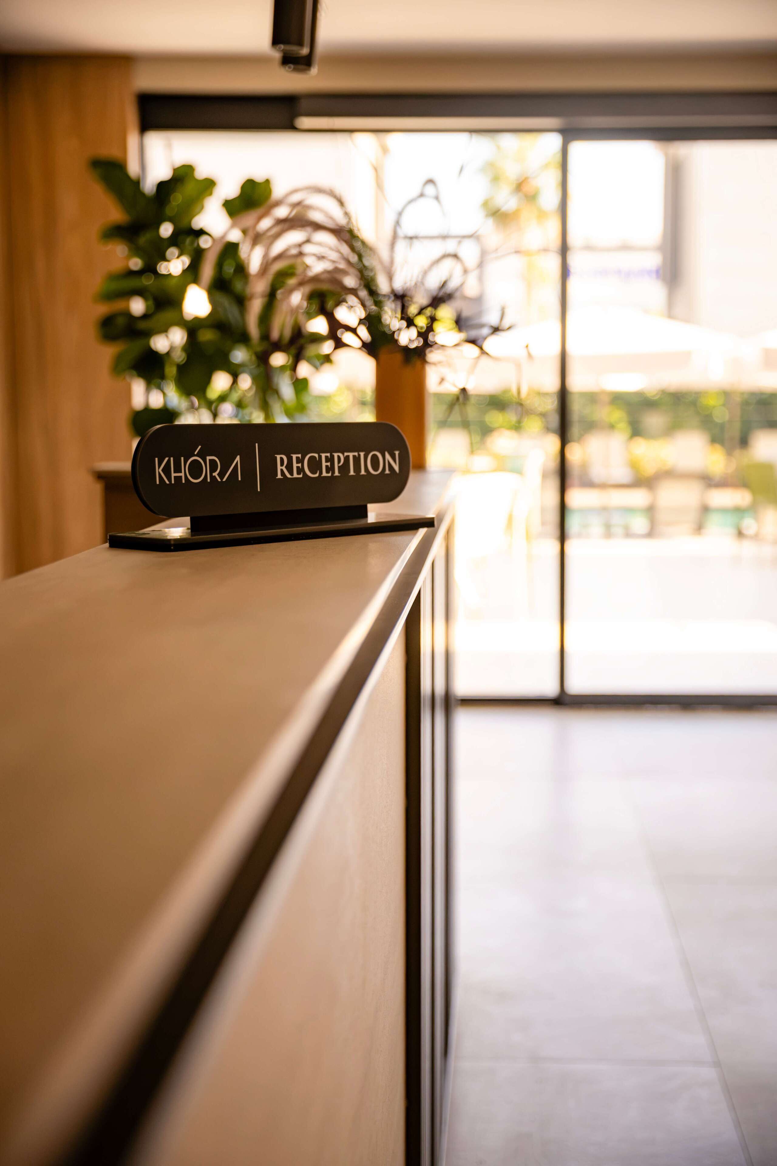 Un taulell de recepció amb un cartell que diu 'KHOIRA | RECEPCIÓ' i una planta en testos al fons.