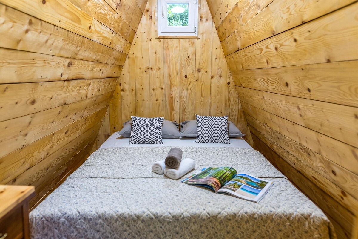 Um pequeno quarto de madeira com teto inclinado, cama com almofadas estampadas, toalha enrolada e uma revista aberta.
