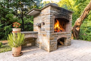 A stone-built outdoor fireplace with a fire burning inside, surrounded by greenery and a potted plant. - Paradise Forest Glamping Rooms (Koper)