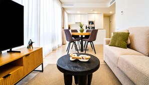 A modern living room with a flat-screen TV, a wooden cabinet, a round black table with pretzel-shaped objects, a beige sofa with a green pillow, and a dining area with chairs and a table. - Unic Serenity Apartment Javea (Javea)