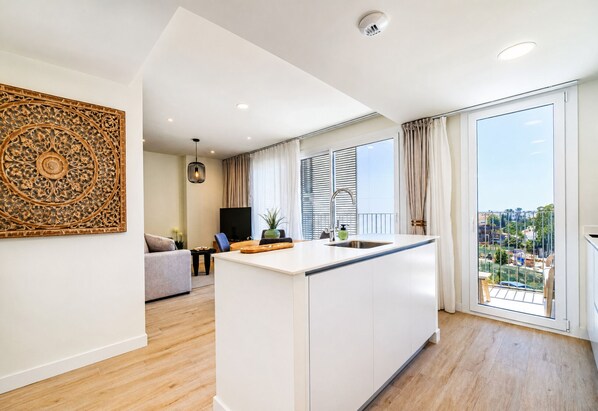Een moderne keuken met een centraal eiland, een zithoek met een bank, en een groot raam met uitzicht op een zwembad en gebouwen.