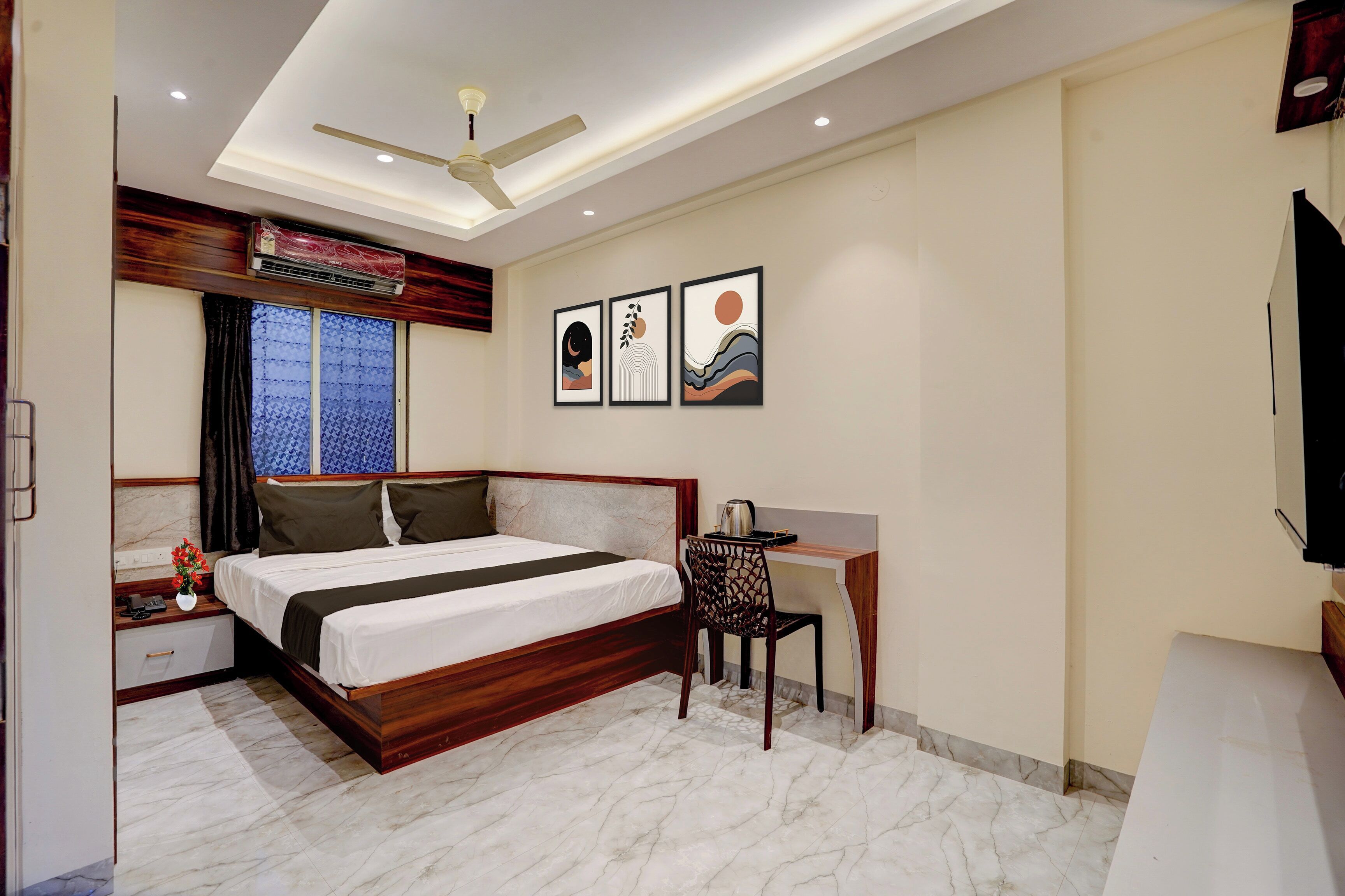 A hotel room with a bed, desk, chair, artwork, and a ceiling fan.